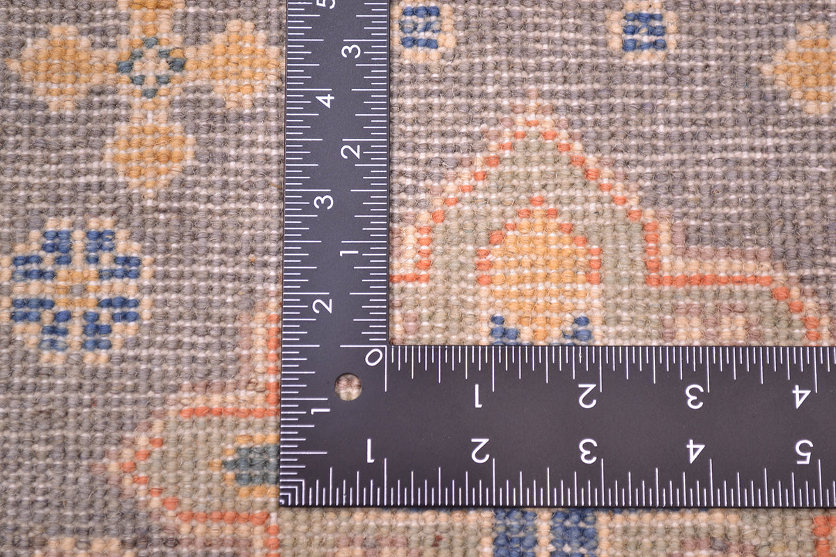 Close-up of the hand-knotted back of a handmade Kazak Strip with a geometric pattern in beige, grey, orange, and blue, shown with a black ruler at the corner to indicate scale.