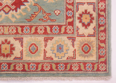 Close-up of a patterned Kazak rug corner showing a cream main border, red narrow borders, and a light blue-green outer edge with fringe.