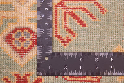 Close-up of the hand-knotted weave of a patterned Sea Green Kazak rug with a dark ruler placed at the corner to show the scale of the knotting, featuring a beige, red, and ochre geometric pattern.