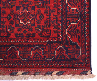 A close-up of an Afghan runner rug corner showing a deep red field with dark navy blue geometric medallions and borders, accented with beige details and fringes with kilim ends.