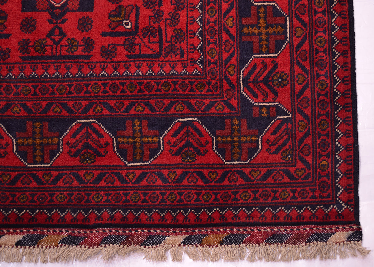 A close-up of a patterned Afghan rug corner showing a deep red field with dark navy geometric motifs, decorative borders, and beige fringe along the bottom edge.