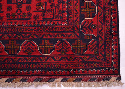 A close-up of a patterned Afghan rug corner showing a deep red field with dark navy geometric motifs, decorative borders, and beige fringe along the bottom edge.