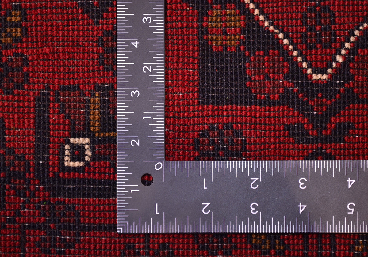 Close-up of the back of a patterned red and black rug with a metal L-square ruler placed over it for scale, showing the textile texture and motif details.