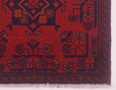 A close-up of corner of a red patterned afghan rug corner with dark navy blue and olive green floral motifs and fringed edges.