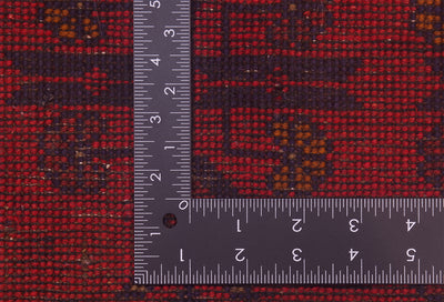 Close-up of back of a red patterned afghan rug with a dark ruler placed on top to show scale, highlighting the textile's weave and design.