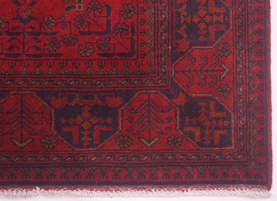 A close-up of a traditional afghan patterned rug corner, showing a deep red field with dark navy blue geometric medallions and borders, accented with small tan details and white fringe along the bottom edge.