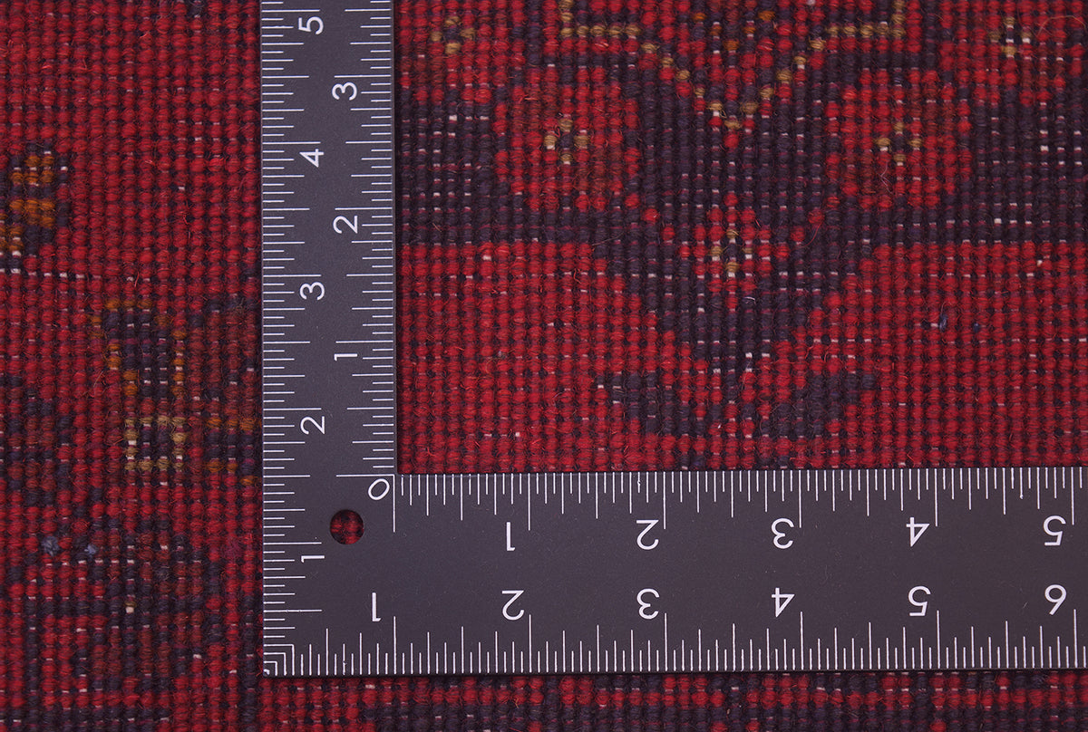 A close-up of back of a red afghan patterned are a rug with a dark floral motif, shown with a metal ruler to indicate scale.