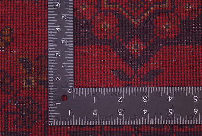 A close-up of back of a red afghan patterned are a rug with a dark floral motif, shown with a metal ruler to indicate scale.