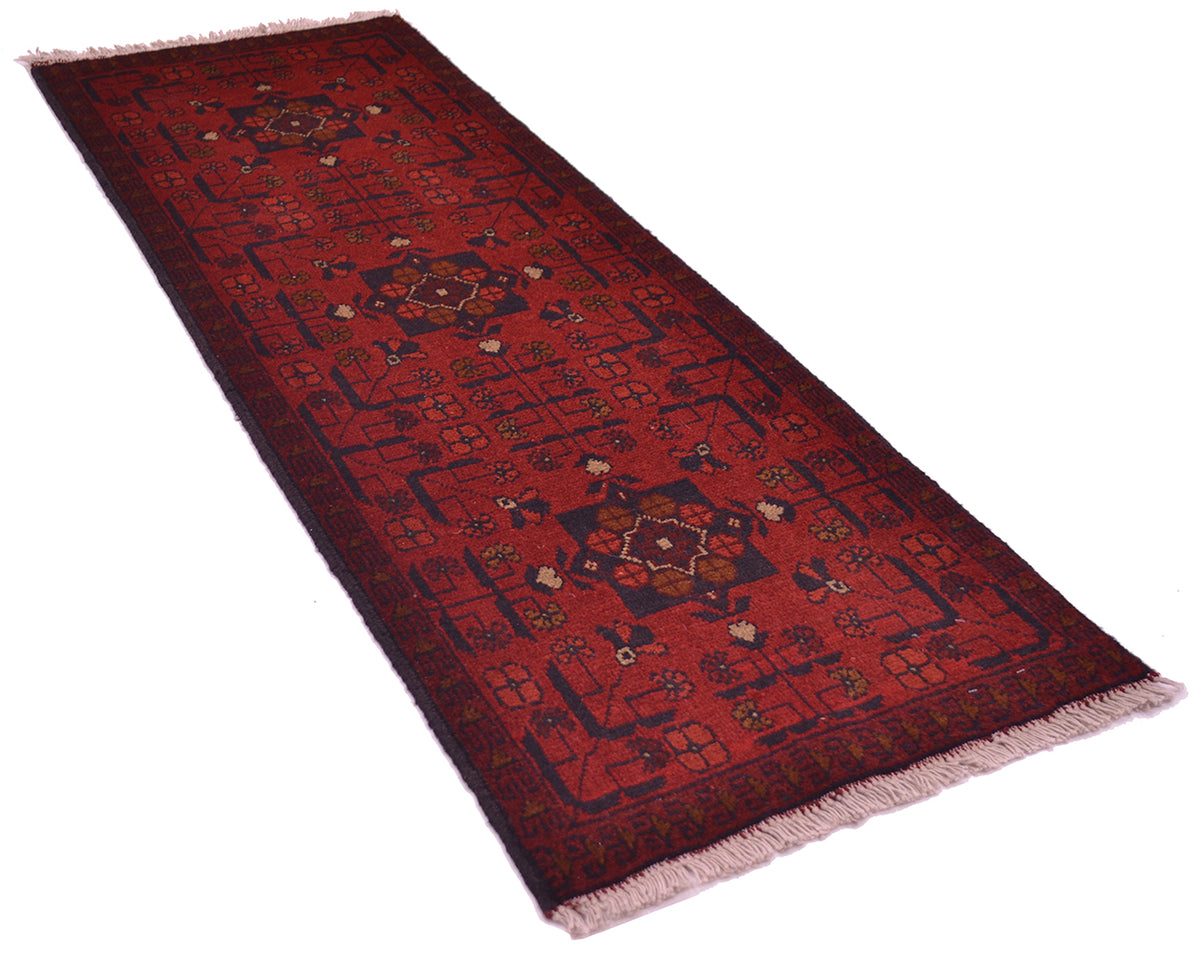 A red afghan runner rug with intricate geometric and floral patterns in dark blue, beige, and brown, finished with fringe on a white background.