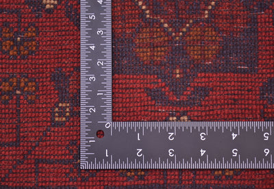 Close-up of back of a patterned with a metal L-square ruler placed on it to show scale. The runner has a dark red base with navy and brown geometric and floral motifs.