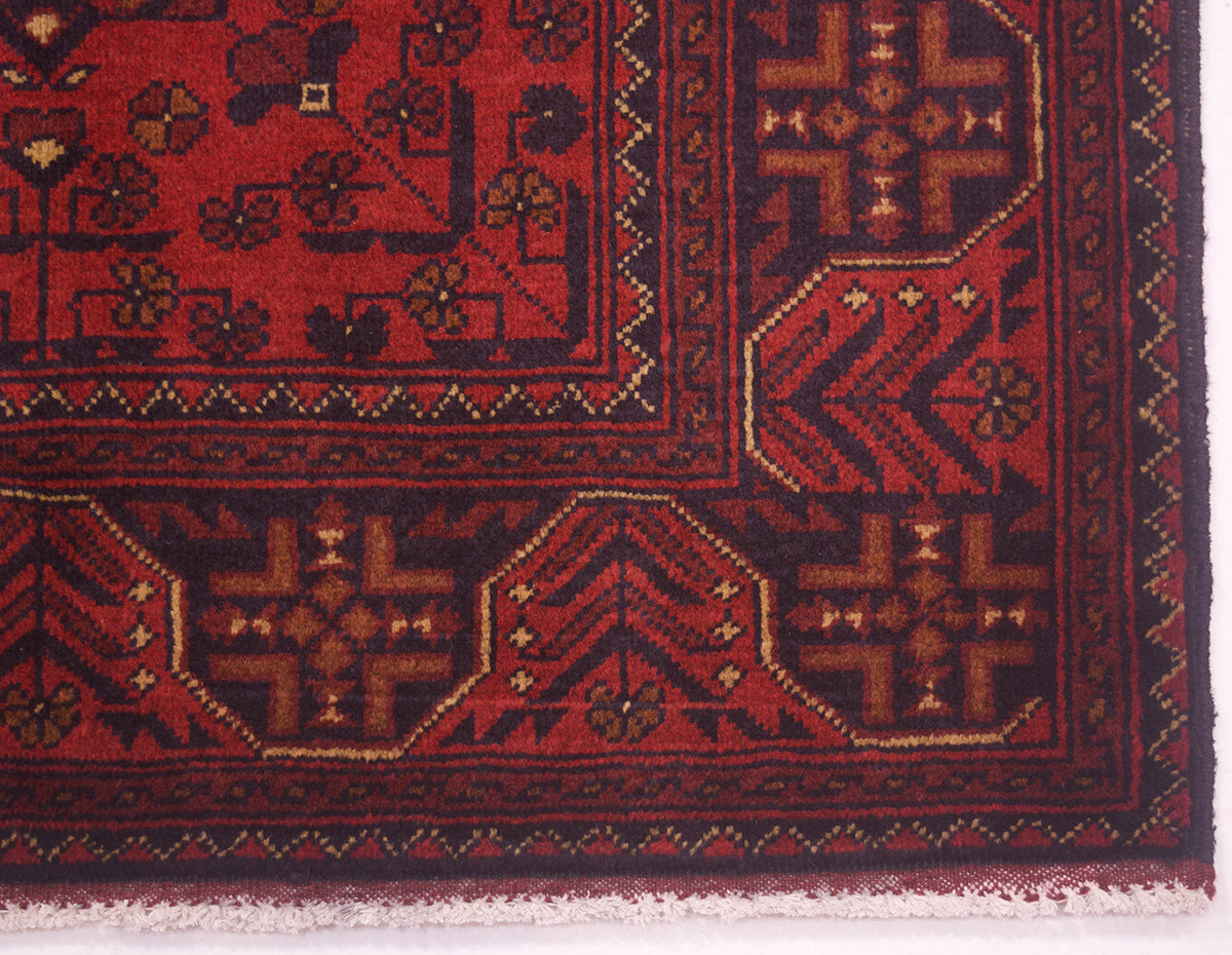 Close-up of a patterned afghan area rug corner showing a deep red field with dark navy borders and geometric floral motifs in brown and beige.