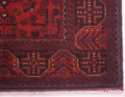 Close-up of a patterned afghan area rug corner showing a deep red field with dark navy borders and geometric floral motifs in brown and beige.