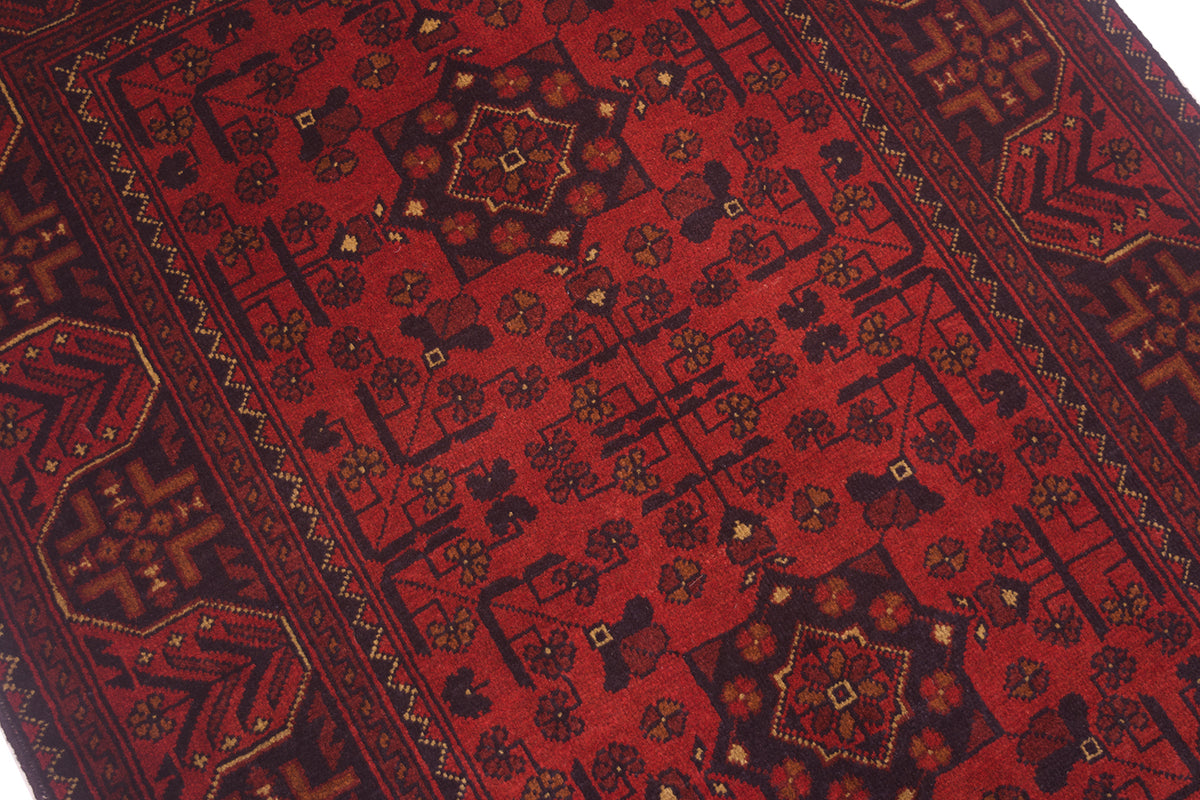 A close-up of a traditional Afghan  patterned rug with a deep red field, dark  borders, and intricate geometric and floral motifs in black, brown, and beige.