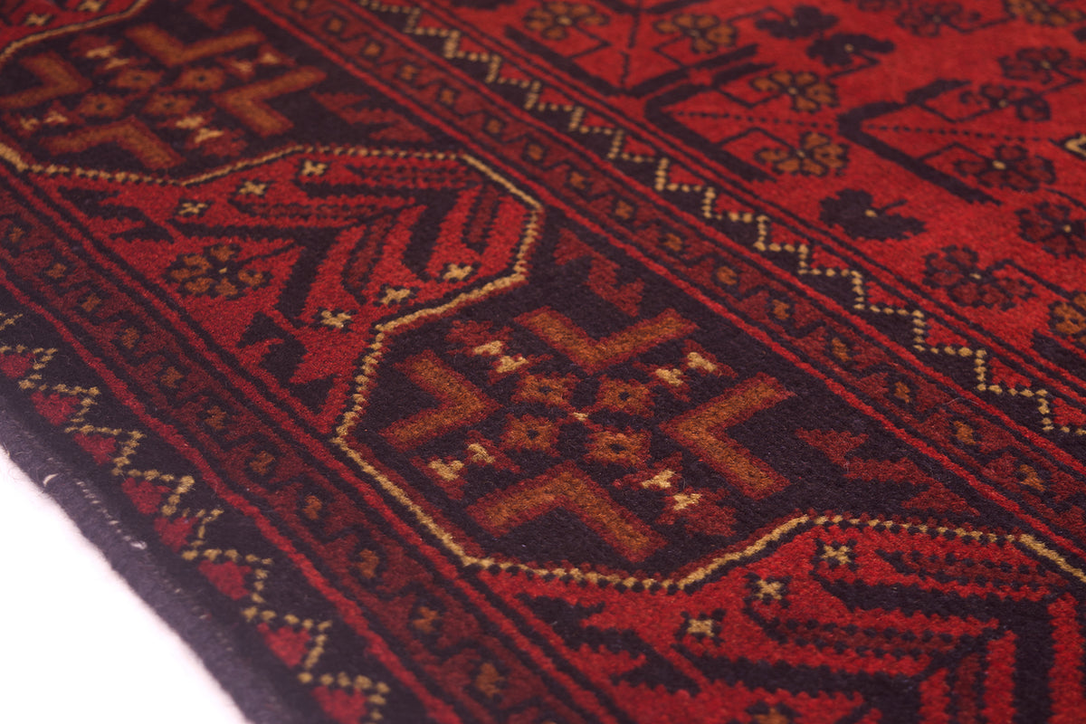 A close-up of a traditional patterned Afghan rug with a deep red field, dark borders, and geometric motifs accented with beige and gold threads.