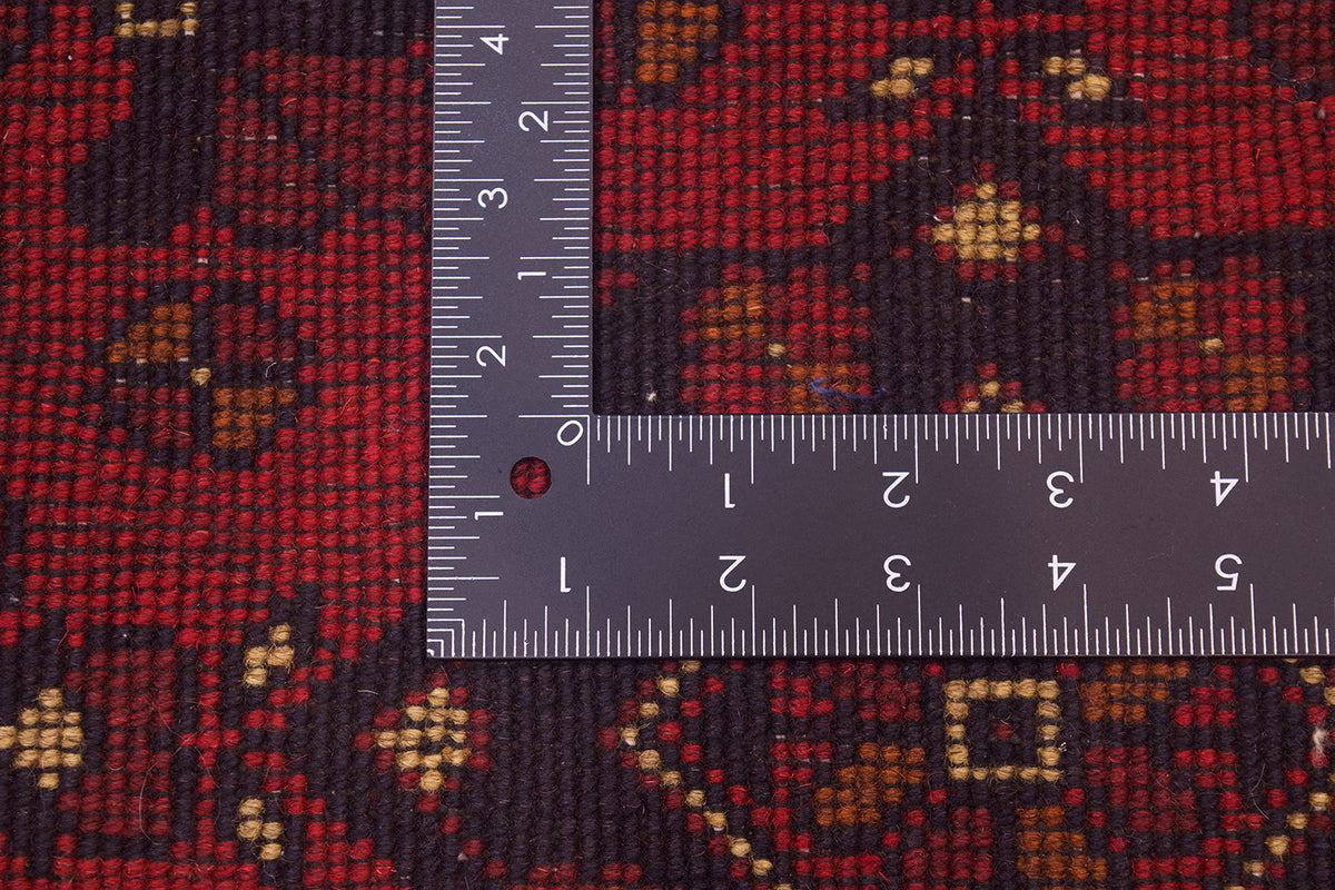 Close-up of back of a patterned afghan area rug with a dark red and black geometric design and small tan accents, shown with a metal ruler to indicate scale.