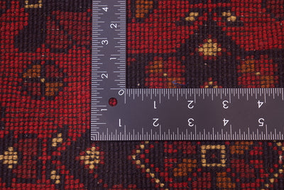 Close-up of back of a patterned afghan area rug with a dark red and black geometric design and small tan accents, shown with a metal ruler to indicate scale.