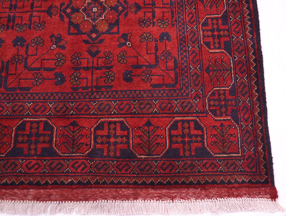 A close-up of a traditional afghan rug corner, showing a deep red field with dark navy blue geometric borders and motifs, finished with white fringe.