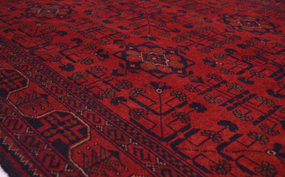 A close-up view of a traditional patterned handmade rug with a deep red field, dark navy geometric borders, and small floral motifs.