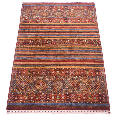 A rectangular hand-woven Khurjeen area rug with a traditional tribal design, featuring horizontal bands of red, blue, brown, and gold geometric patterns, finished with a fringed edge.