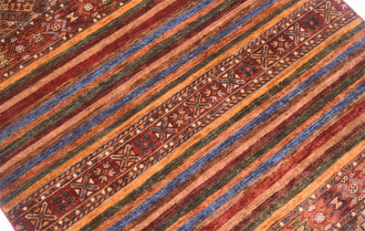 A close-up of a patterned Khurjeen rug with diagonal stripes in rust red, orange, blue, green, and brown, plus geometric border motifs.