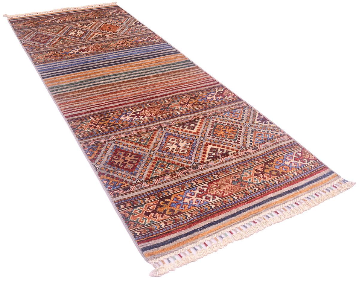 A long rectangular Khurjeen runner with a complex geometric tribal pattern in shades of rust red, blue, beige, and brown, finished with cream fringe on both ends.