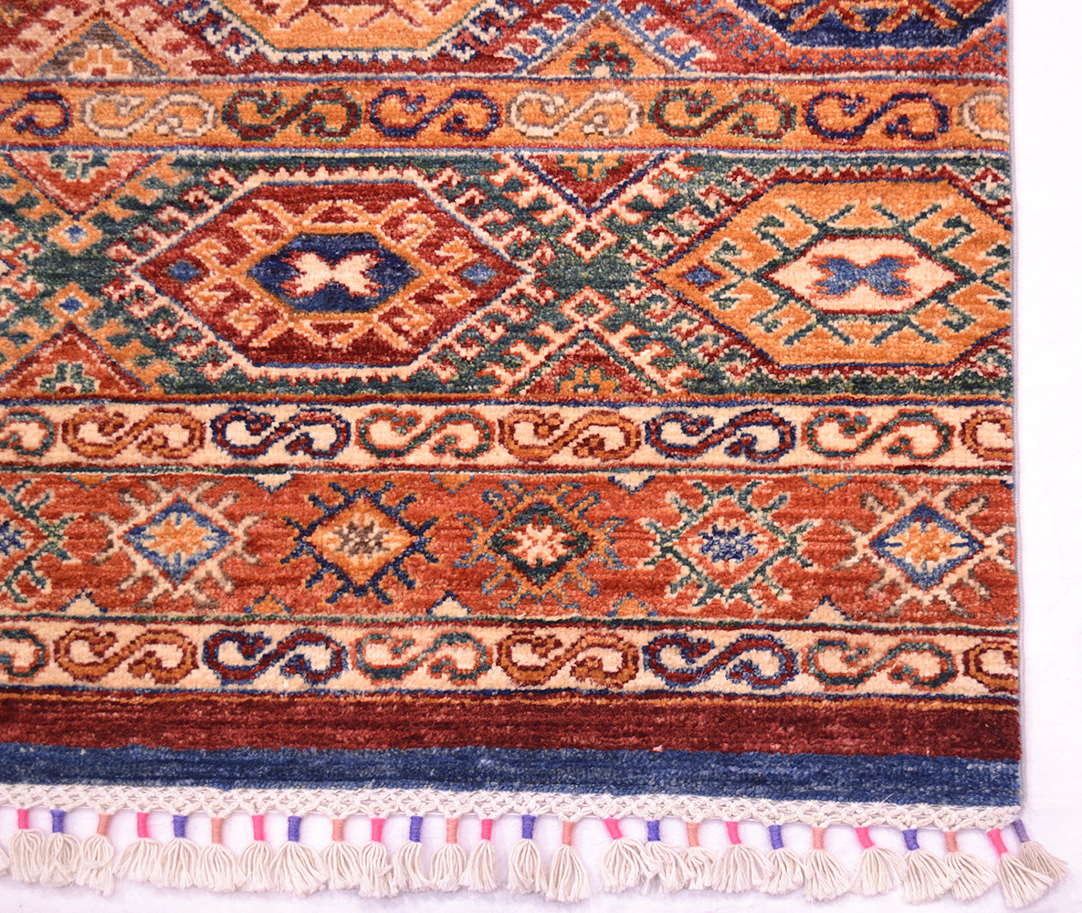 A close-up of a corner of patterned Afghan Khorjin rug showing geometric motifs in rust orange, navy blue, beige, and green, with cream fringe along the bottom edge.