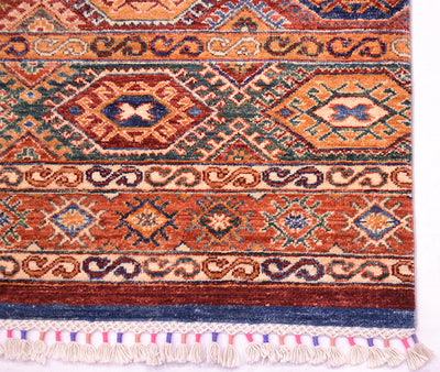 A close-up of a corner of patterned Afghan Khorjin rug showing geometric motifs in rust orange, navy blue, beige, and green, with cream fringe along the bottom edge.