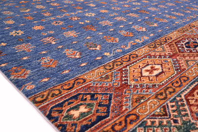 A close-up of a traditional patterned Khurjeen carpet with a blue field, orange and beige floral motifs, and a detailed geometric border in rust, navy, and cream.