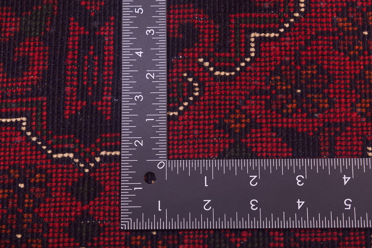 Close-up of back of a red patterned afghan rug with a dark navy/black base and cream geometric accents, shown with a metal L-square ruler for scale.