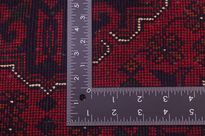 Close-up of back of a red patterned afghan rug with a dark navy/black base and cream geometric accents, shown with a metal L-square ruler for scale.