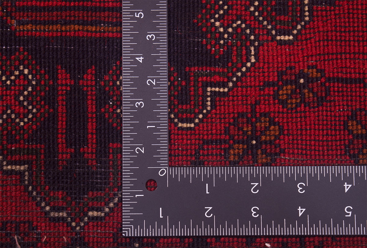 A close-up of back of a red afghan rug with dark navy and orange/brown motifs, shown with a metal ruler to indicate scale.