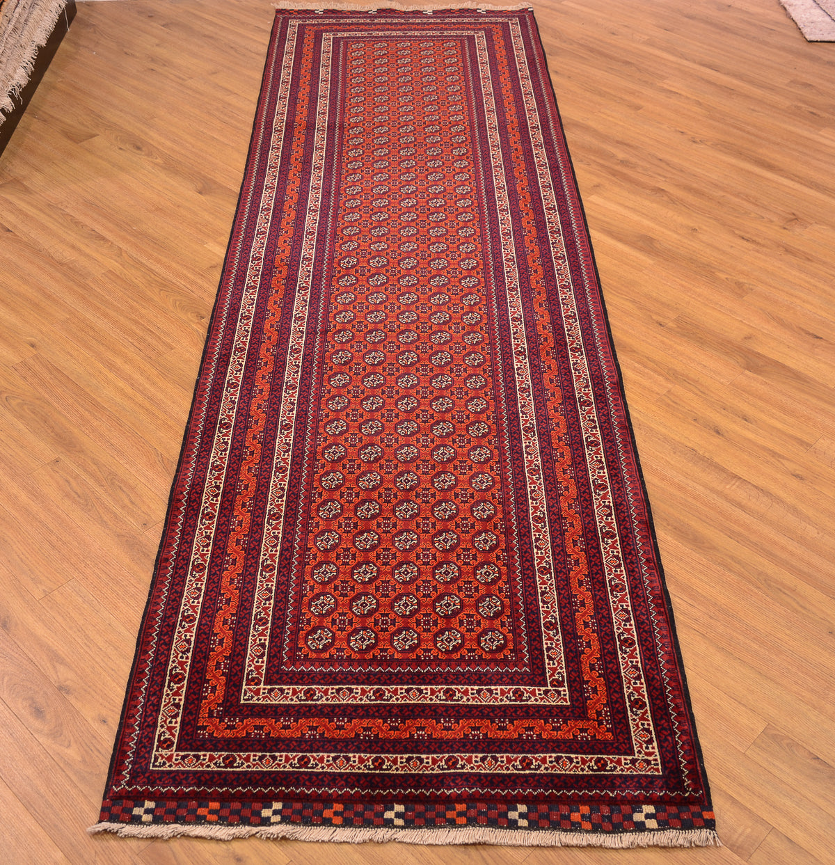 A long Afghan runner rug with an intricate traditional pattern, shown laid out on a light wooden floor. The rug features a deep red center, dark navy blue borders, and accents of orange, beige, and white in the geometric and floral motifs.