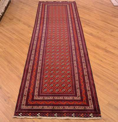 A long Afghan runner rug with an intricate traditional pattern, shown laid out on a light wooden floor. The rug features a deep red center, dark navy blue borders, and accents of orange, beige, and white in the geometric and floral motifs.
