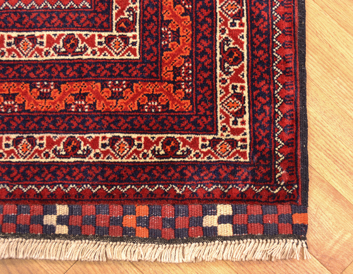 A close-up of a traditional afghan patterned rug corner on a light wood floor, showing red, navy, beige, and orange motifs with cream fringe.