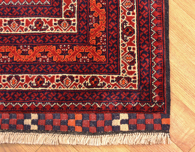 A close-up of a traditional afghan patterned rug corner on a light wood floor, showing red, navy, beige, and orange motifs with cream fringe.