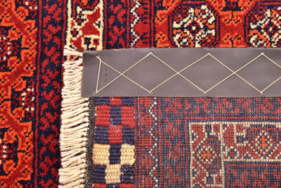 Close-up of back of a patterned Afghan runner rug corner showing cream fringe, a gray strapping attached to edge, and red, navy, and beige woven textile details.