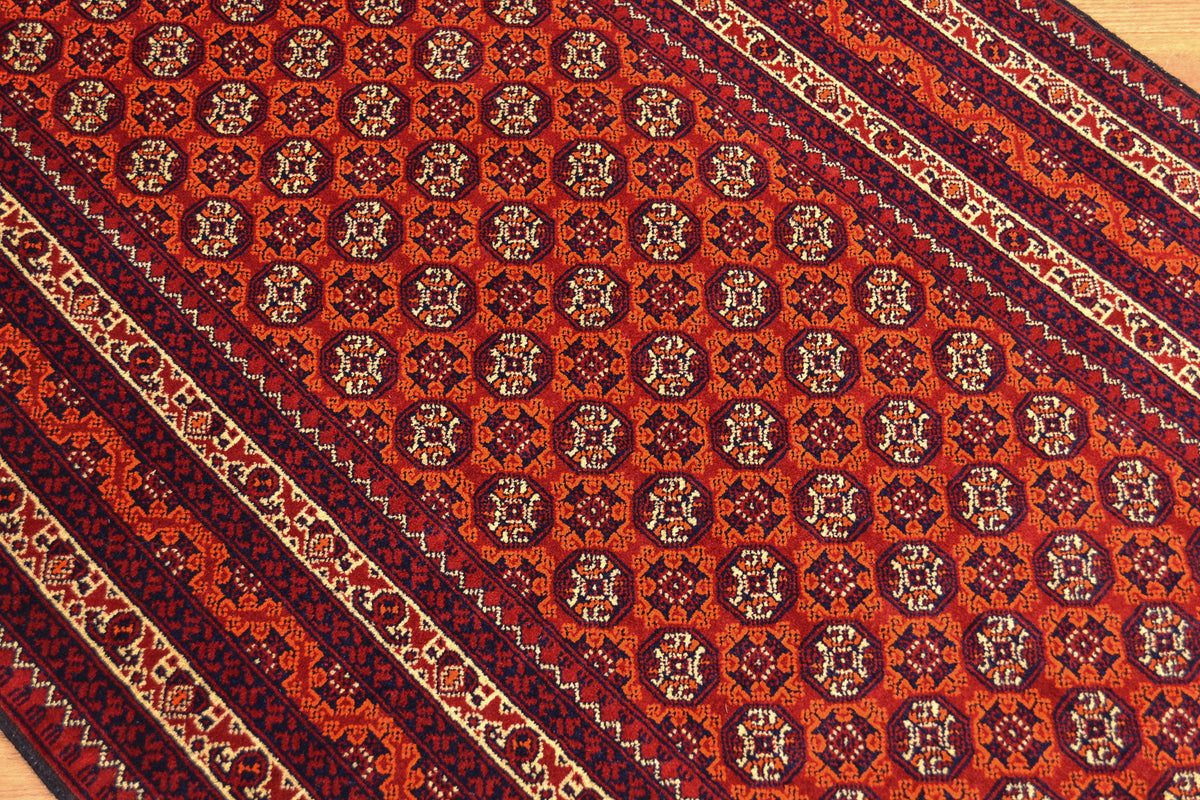 A close-up of a patterned Afghan runner rug with a warm rust-red field, repeating medallion motifs, and dark navy and beige borders.