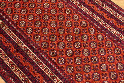 A close-up of a patterned Afghan runner rug with a warm rust-red field, repeating medallion motifs, and dark navy and beige borders.