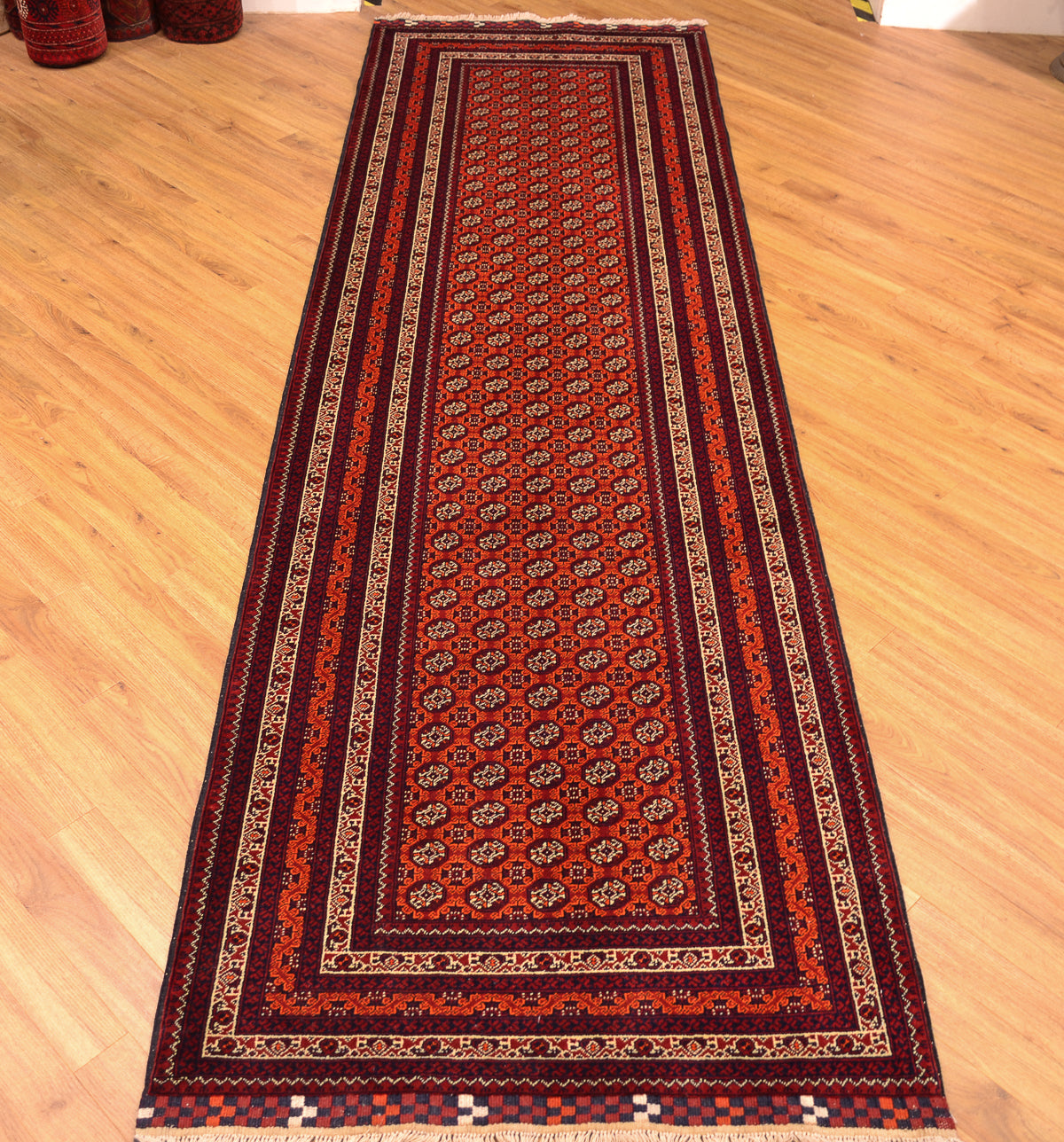 A long rectangular area runner rug with an intricate traditional pattern, laid out on a light wooden floor. The rug features a deep red center, dark maroon borders, and cream and beige geometric accents.
