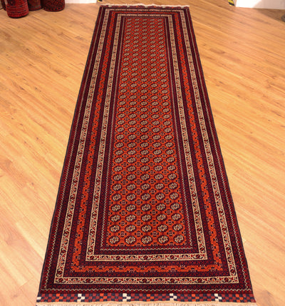 A long rectangular area runner rug with an intricate traditional pattern, laid out on a light wooden floor. The rug features a deep red center, dark maroon borders, and cream and beige geometric accents.