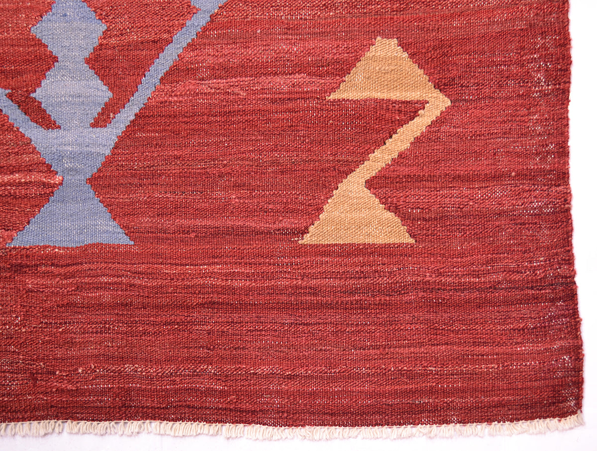 Close-up of a handwoven red Labijar Kilim rug with a rust red field, a large light blue geometric motif on the left, a tan zigzag shape on the right, and a short fringe along the bottom edge.