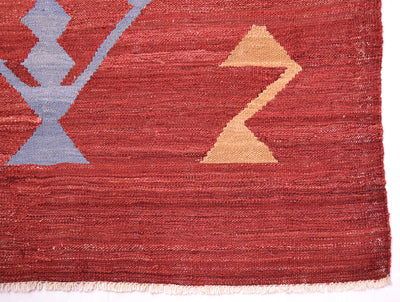 Close-up of a handwoven red Labijar Kilim rug with a rust red field, a large light blue geometric motif on the left, a tan zigzag shape on the right, and a short fringe along the bottom edge.