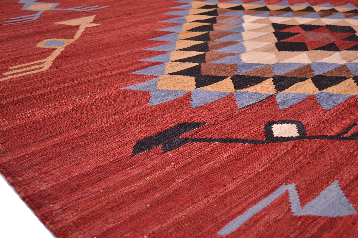 A close-up of a handwoven red Labijar Kilim rug with a textured red field, featuring geometric tribal patterns in beige, blue, black, and brown.