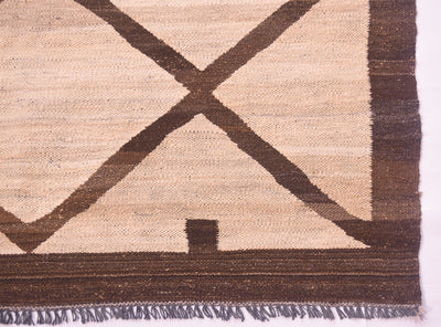 Close-up of a tribal style handwoven Afghan Kilim showing a beige natural-wool base with a dark brown geometric pattern, including a large X shape, a vertical border, and fringe along the bottom edge.