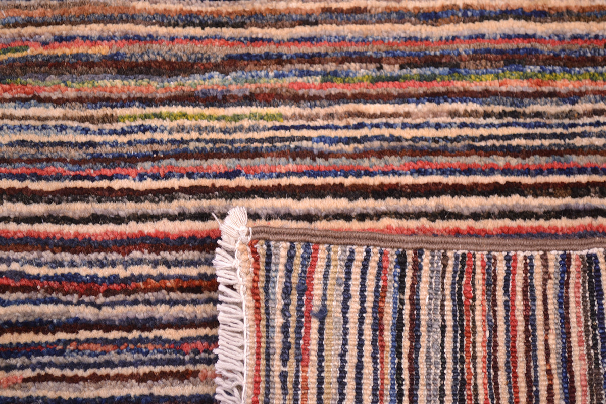 Close-up of a corner turned up to show the weave of a handmade stripy Gabbeh rug with multicoloured horizontal and vertical bands in beige, brown, blue, red, and cream, showing a fringed edge.