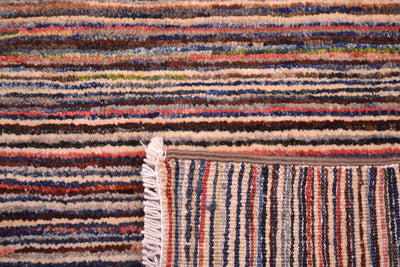 Close-up of a corner turned up to show the weave of a handmade stripy Gabbeh rug with multicoloured horizontal and vertical bands in beige, brown, blue, red, and cream, showing a fringed edge.
