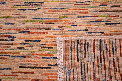 Close-up of a turned over corner of an Afghan Gabbeh carpet with a multicolour abstract pattern of horizontal and vertical stripes in beige, orange, blue, red, and green, featuring a cream-coloured fringed edge.