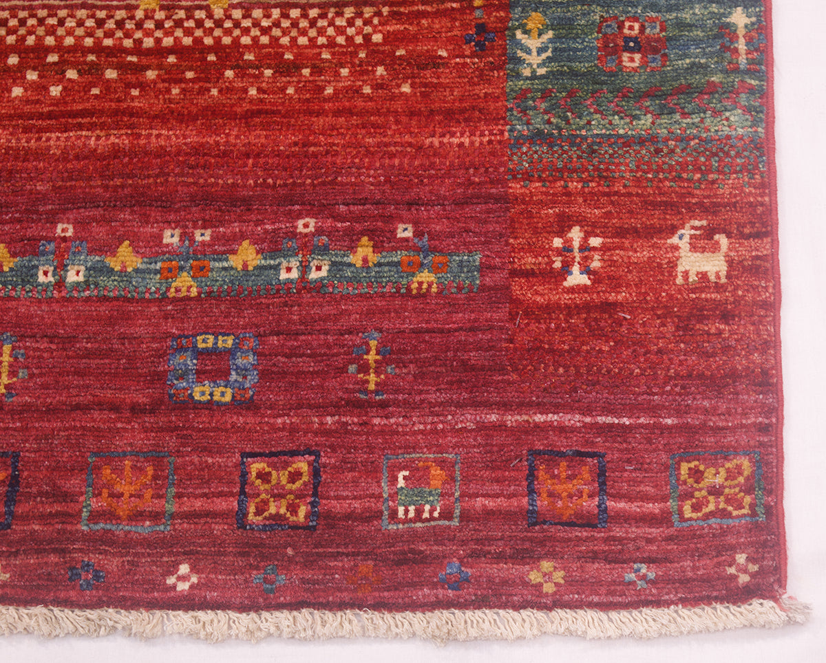 A close-up of a corner of a handwoven colourful Afghan Loribaft rug with a deep red base and geometric patterns in blue, green, orange, and cream, featuring a fringed edge.