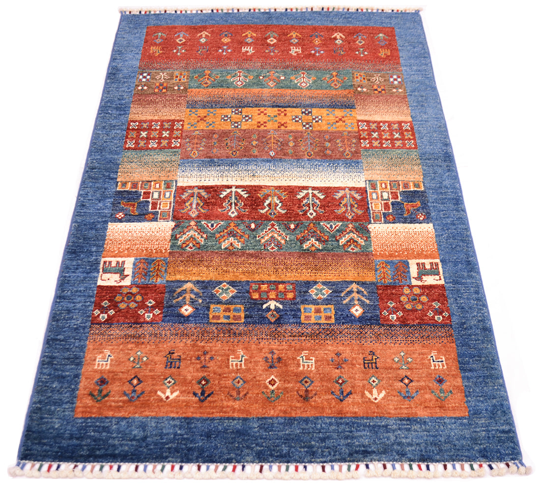 A rectangular hand-knotted Afghan Loribaft area rug with a blue border and multiple horizontal bands of geometric and floral patterns in rust red, orange, beige, and teal, finished with white fringe on the top and bottom.