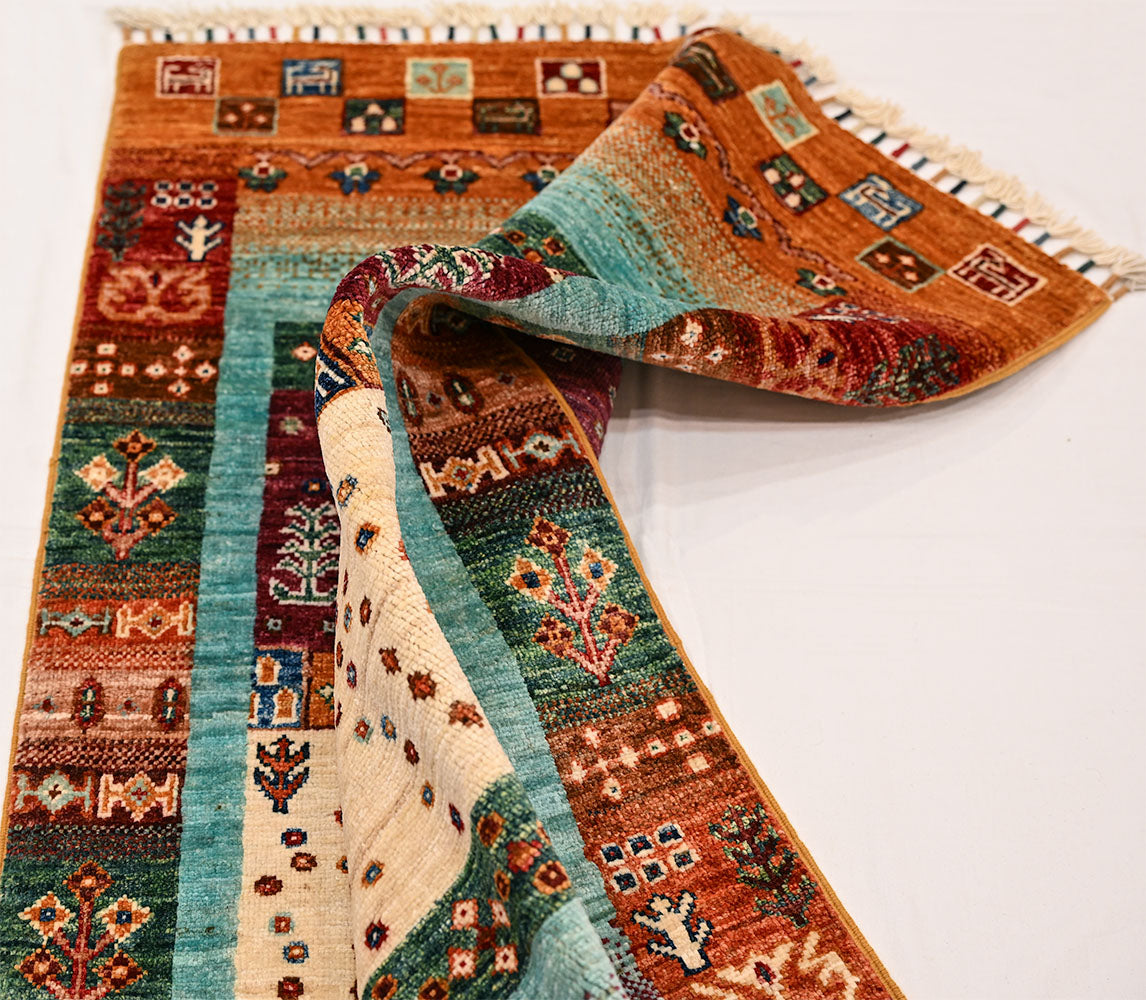 Loribaft Gabbeh rug draped with turquoise and rust bands, multicolour patterns, and fringed edge along the top of the picture.